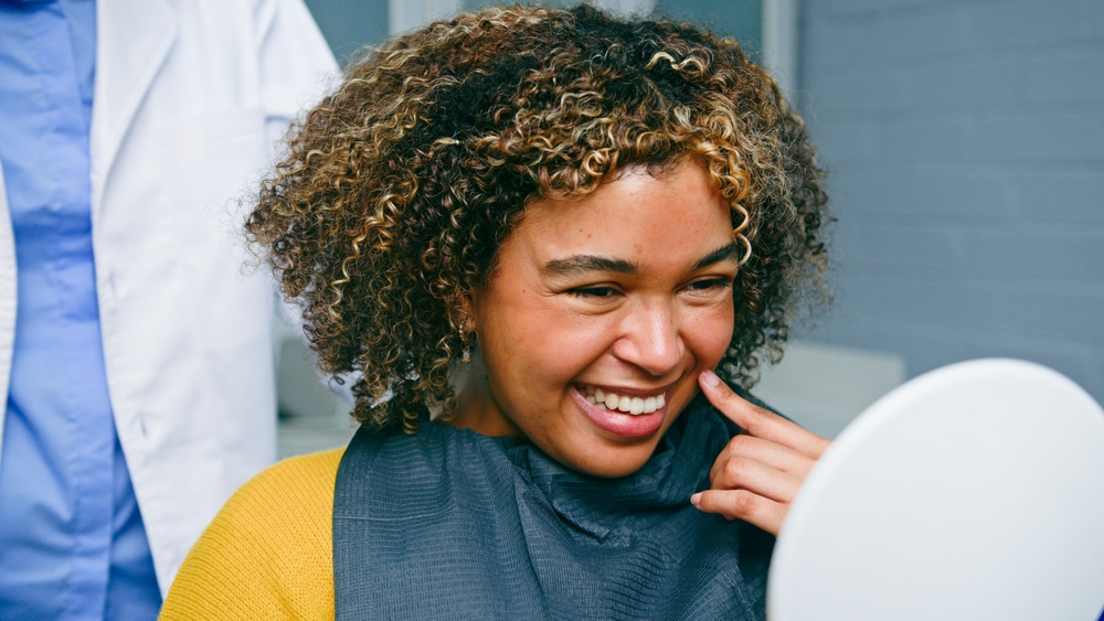 A woman with curly hair smiles as she looks at her reflection in a handheld mirror. She has on a dental bib and points to her teeth. Beside her, a person in a white coat is partly visible in what looks like a dental clinic.