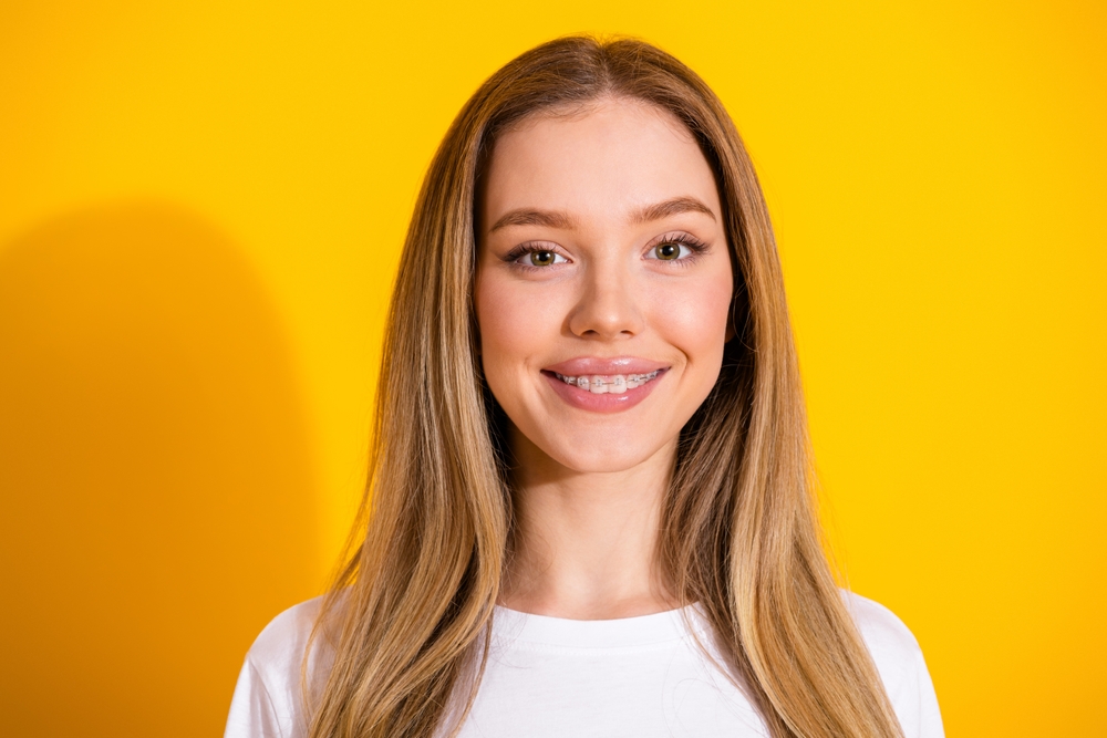 A young woman with long, straight blonde hair and braces smiles at the camera. She is wearing a white shirt and stands in front of a vibrant yellow background.