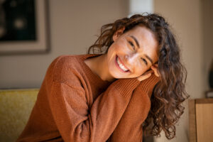 A woman with curly brown hair, wearing a rust-colored sweater, smiles warmly while leaning her head on her hands. She sits indoors in soft natural light with a blurred background.