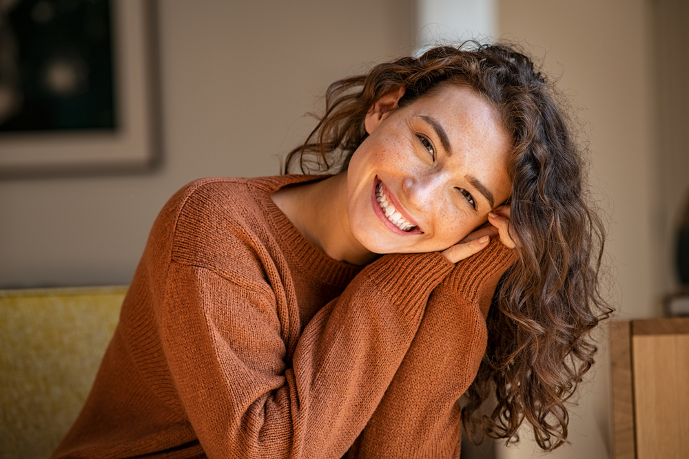 A woman with curly brown hair, wearing a rust-colored sweater, smiles warmly while leaning her head on her hands. She sits indoors in soft natural light with a blurred background.