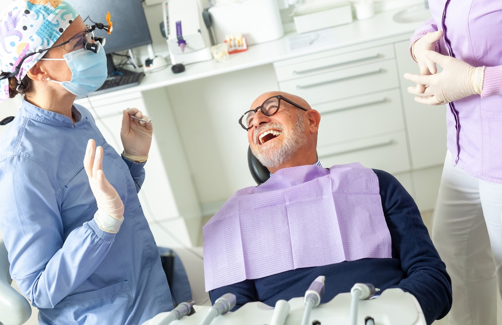 A bald man wearing glasses sits in a dental chair, laughing. A dentist in a blue coat, mask, and colorful cap talks to him, while another dental staff member in a lavender coat stands nearby. Dental tools are visible in the foreground.