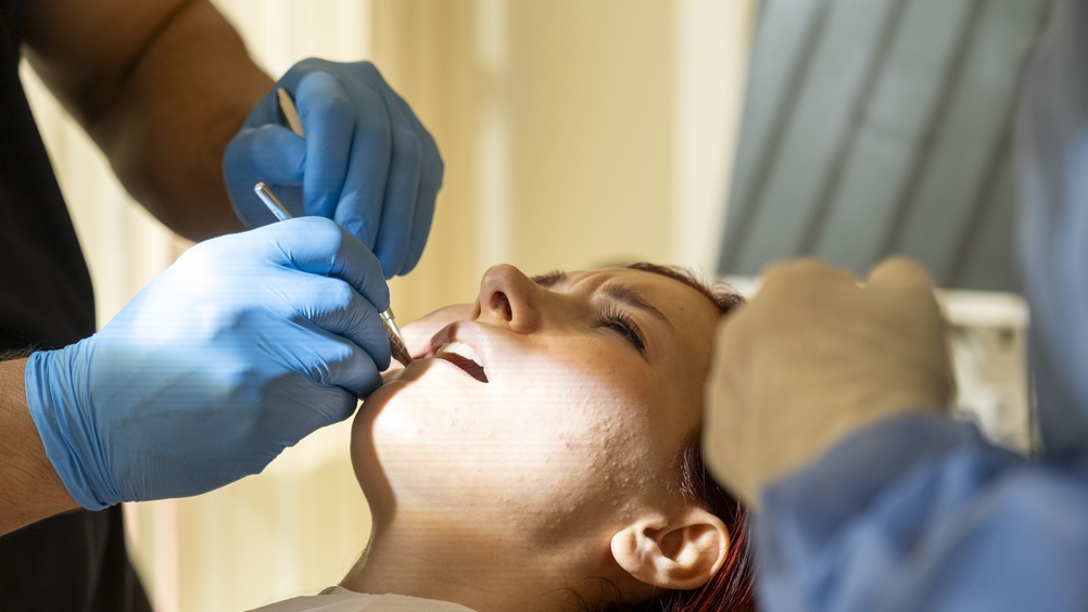 A person lies in a dental chair with their mouth open while two gloved dentists use dental instruments to examine or treat their teeth under bright lighting.