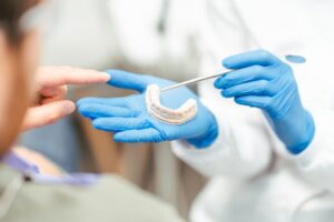 A dentist wearing blue gloves holds a dental mold and points with tweezers, while a patient points at the model during a consultation. Attention is on the hands and the dental model.