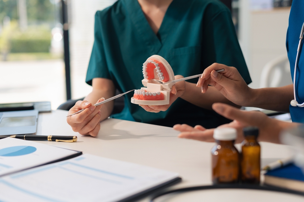 Two people in scrubs hold a dental model and use dental tools to explain dental care. Bottles, papers, and a pen are on the table, suggesting a dental consultation or educational session.